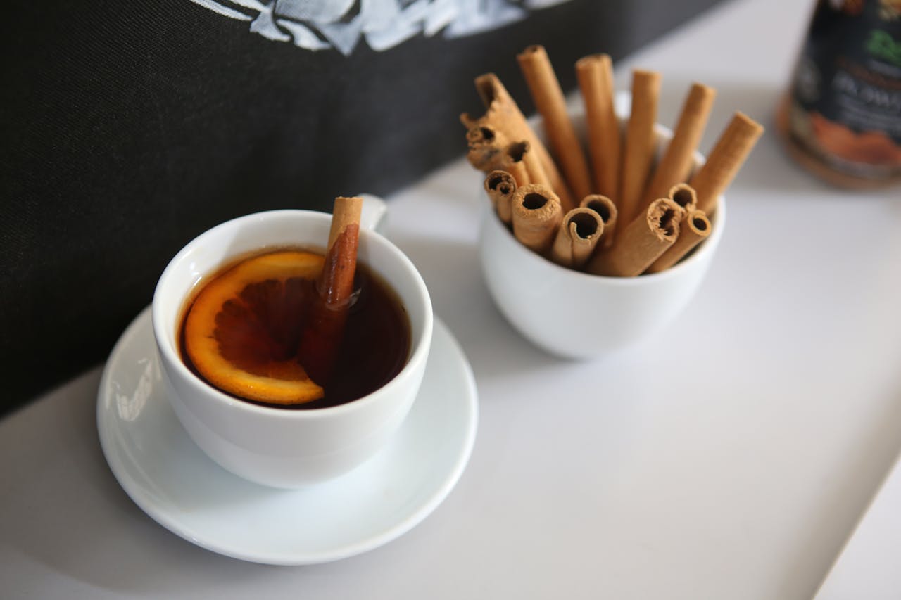 our-experience A white cup of cinnamon-flavored tea with a citrus slice, accompanied by cinnamon sticks.